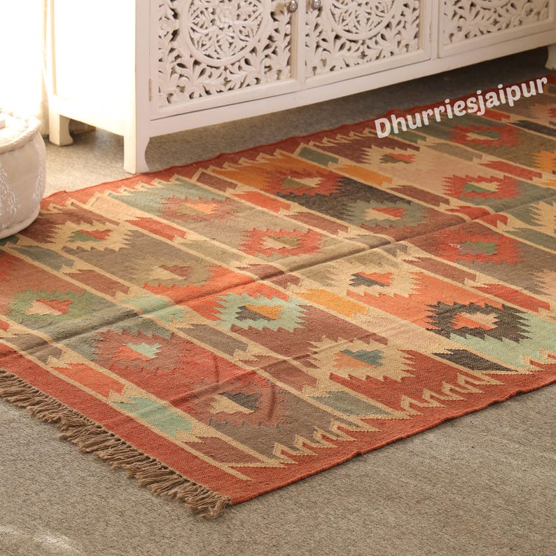 May include: A rectangular rug with a geometric pattern in shades of red, orange, brown, and beige. The rug has a fringed edge and the text "Dhurriesjaipur" is visible in the upper right corner. The rug is placed on a gray surface.