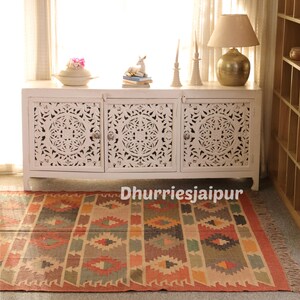 May include: A white wooden cabinet with carved floral designs on the doors, topped with decorative items. A colorful patterned rug is in front of the cabinet. The text "Dhurriesjaipur" is at the bottom of the image.