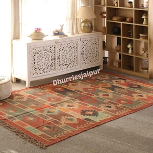 May include: A rectangular, patterned rug with a fringed edge. The rug features a geometric design in shades of red, orange, brown, and green. The rug is placed on a gray floor in front of a white cabinet.