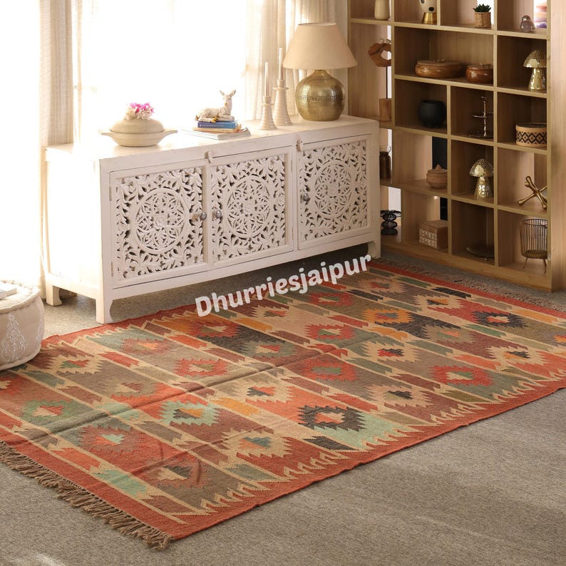 May include: A rectangular, patterned rug with a fringed edge. The rug features a geometric design in shades of red, orange, brown, and green. The rug is placed on a gray floor in front of a white cabinet.