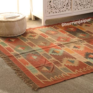 May include: A rectangular, patterned rug with a fringed edge. The rug features a geometric design in shades of red, orange, brown, and beige. The rug is placed on a gray surface, with a beige ottoman and white cabinet in the background.
