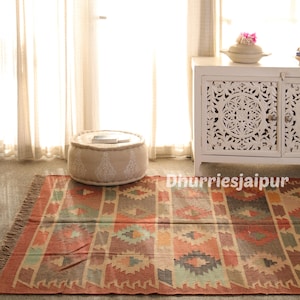 May include: A colorful, patterned rug with a fringed edge. The rug features geometric designs in shades of red, orange, green, and blue. A beige ottoman with white embroidered details sits on the rug. A white cabinet is in the background.