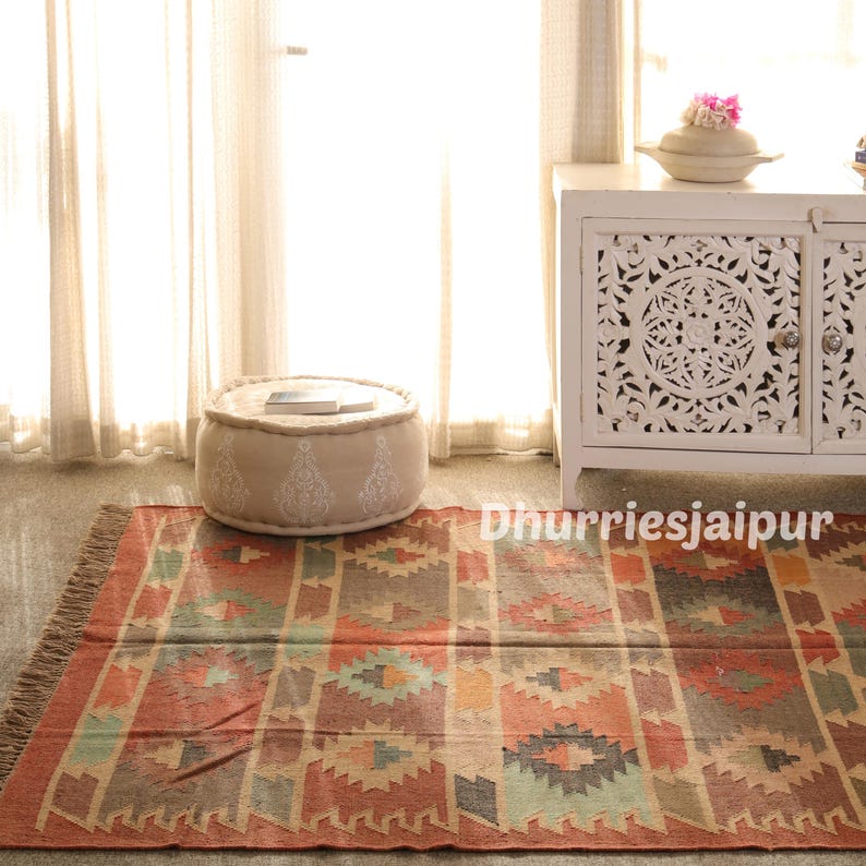 May include: A colorful, patterned rug with a fringed edge. The rug features geometric designs in shades of red, orange, green, and blue. A beige ottoman with white embroidered details sits on the rug. A white cabinet is in the background.