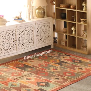 May include: A colorful, patterned rug with a geometric design in shades of orange, brown, green, and blue. The rug is rectangular and appears to be made of woven fabric. The text "Dhurriesjaipur" is visible on the rug.