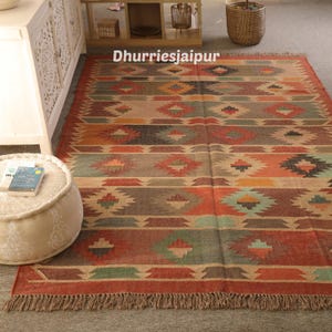 May include: A rectangular area rug with a geometric pattern in shades of red, orange, green, and brown. The rug has a fringed edge and the text "Dhurriesjaipur" is visible on the rug.