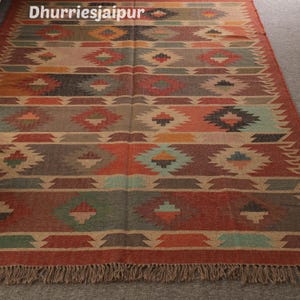 May include: A rectangular rug with a geometric pattern in shades of red, orange, green, and brown. The rug has a fringed edge and the text "Dhurriesjaipur" at the top. The design features diamond and zigzag shapes.