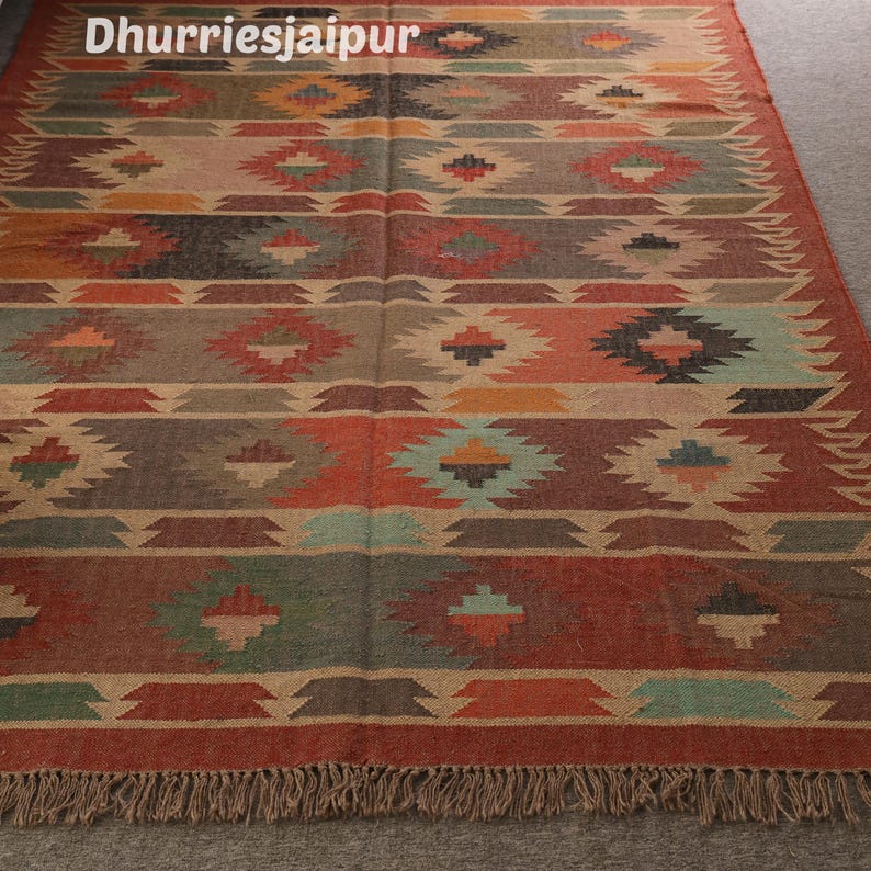 May include: A rectangular rug with a geometric pattern in shades of red, orange, green, and brown. The rug has a fringed edge and the text "Dhurriesjaipur" at the top. The design features diamond and zigzag shapes.