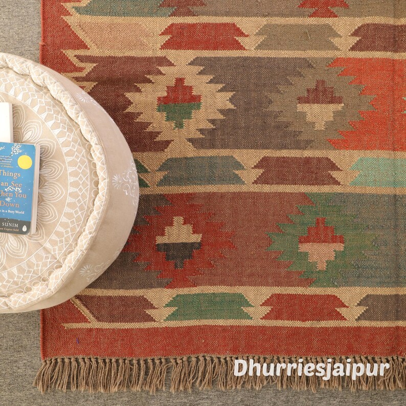 May include: A rectangular, patterned rug with a fringe. The rug features geometric designs in shades of red, brown, green, and beige. A round, beige ottoman with a book on top is in the upper left corner. The text "Dhurriesjaipur" is at the bottom.