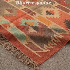 May include: A colorful area rug with a geometric pattern in shades of red, orange, brown, and teal. The rug has a fringed edge and the text "Dhurriesjaipur" in the upper left corner.