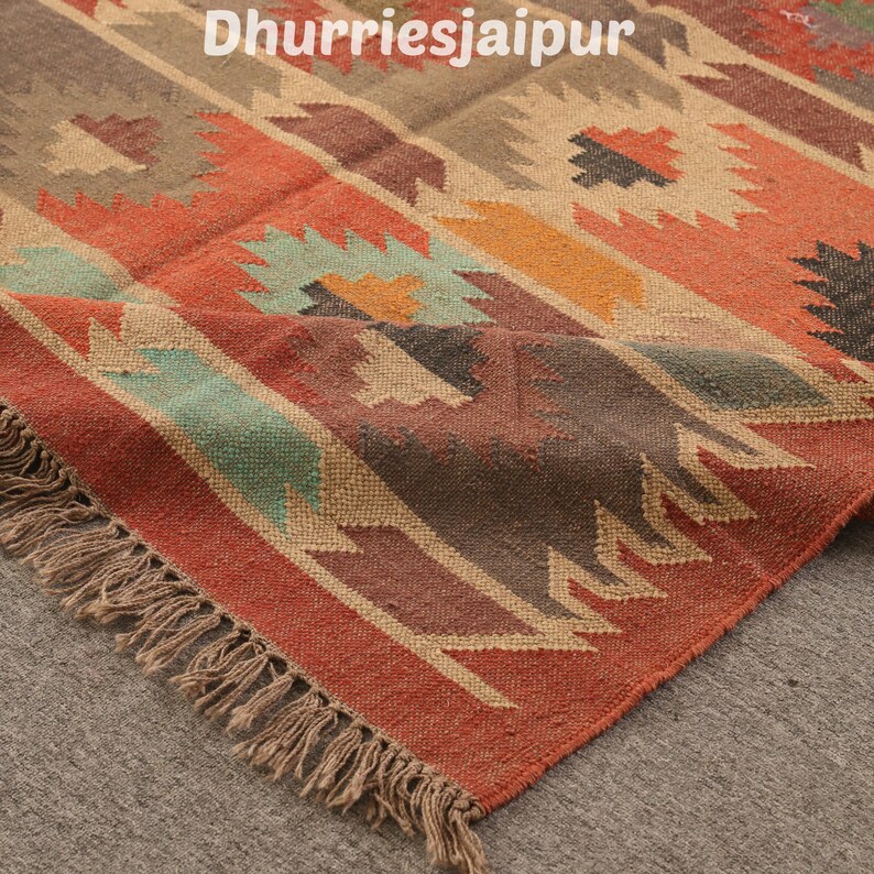 May include: A colorful area rug with a geometric pattern in shades of red, orange, brown, and teal. The rug has a fringed edge and the text "Dhurriesjaipur" in the upper left corner.