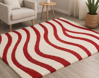 May include: A rectangular area rug featuring a repeating pattern of thick, wavy red stripes set against a cream-colored background. The rug has a textured, plush pile surface and covers a portion of the light-colored wooden floor in the room.