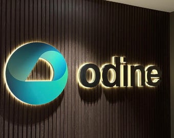 May include: Illuminated sign with the word "odine" in a modern font, mounted on a dark wood-paneled wall. The sign features a teal and blue circular logo with a gradient effect. The letters are outlined with a warm light.