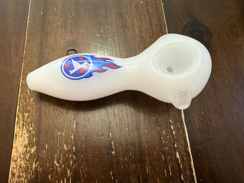 May include: White glass pipe with a rounded bowl and a stem. The pipe features a blue and red flame design with a circular emblem. The emblem has a white bird and three red stars.