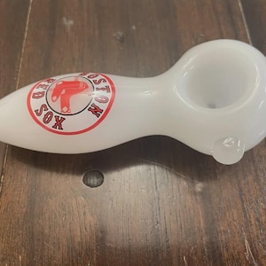 May include: White glass pipe with a rounded bowl and a stem. The pipe features a circular logo with the words "BOSTON RED SOX" around a red sock graphic. The pipe is designed for smoking.