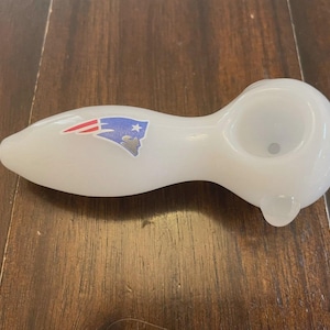 May include: A white glass pipe with a curved design. The pipe features a bowl, a stem, and a mouthpiece. A sticker of the New England Patriots logo is on the side. The pipe is on a wooden surface.