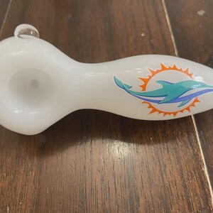 May include: White glass pipe with a rounded bowl and a mouthpiece. The pipe features a teal and orange dolphin logo on the side. A small, clear glass bubble is on top of the bowl.