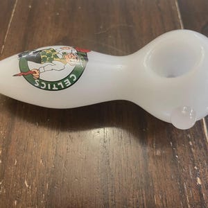 May include: White glass pipe with a rounded bowl and a stem. The pipe features a green and white Celtics logo on the side. The pipe is designed for smoking and rests on a wooden surface.