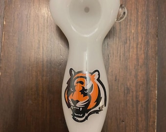 May include: White glass pipe with a tiger logo in orange, black, and white. The pipe has a rounded bowl and a long, tapered stem. A small glass handle is attached to the side of the bowl.