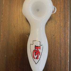 May include: White glass pipe with a rounded bowl and a tapered stem. The stem features a red and black logo with the letters "KC" inside a white arrowhead shape. The pipe is designed for smoking.