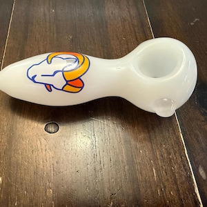 May include: White glass pipe with a bowl and a mouthpiece. The pipe features a colorful ram logo in blue, orange, and yellow. The pipe is smooth and sits on a dark wooden surface.
