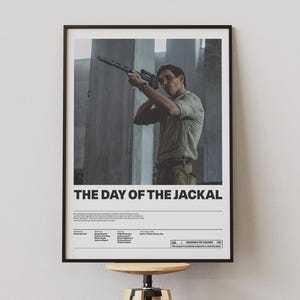 Può includere: Un poster incorniciato del film "Il giorno dello sciacallo". L'immagine mostra un uomo che punta un fucile in un ambiente di cemento. Il titolo è in testo bianco in basso. Il poster è su uno sgabello di legno.