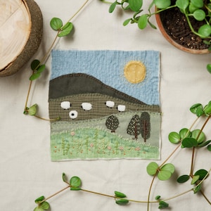 May include: A textile art piece depicting a pastoral scene. The artwork features a blue sky, a yellow sun, a green hill with white sheep, and green trees. The piece is made of various fabrics and textures, creating a handcrafted look.