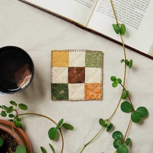 May include: A square, quilted coaster with a patchwork design. The coaster features nine squares in various colors: yellow, brown, green, and orange, with floral and leaf patterns. A cup of tea and a book are also in the image.