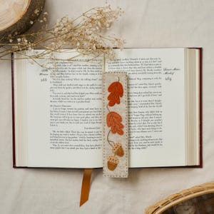 May include: A handmade felt bookmark featuring autumn leaves and acorns in shades of orange and brown. The bookmark is placed in an open book, with the page numbers 268 and 269 visible. A leather bookmark is also visible.
