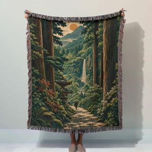May include: A woven throw blanket featuring a detailed landscape scene with tall trees, a waterfall, a distant mountain, and a golden sun. A person walks along a path near a structure. The blanket has a fringed edge and a dark, earthy colour palette.