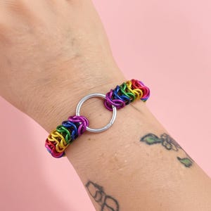 May include: A rainbow-colored chainmail bracelet with a silver-colored circular clasp. The bracelet is made of small, brightly colored metal rings in a rainbow pattern. The bracelet is shown on a wrist against a pink background.