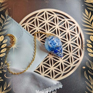 May include: A gold-colored chain with a blue and white pendant. The pendant is attached to a gold chain and rests on a light blue cloth. A gold crescent moon and star are also visible. The background features a geometric design and the text "Maya et Cyal."