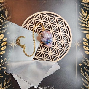 May include: A gold-colored chain necklace with a pendant featuring pink and blue stones. The necklace is displayed with a white pouch and a gold crescent moon charm. The background includes a wooden geometric design and the text "Maya et Cyal."