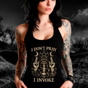 May include: Black halter top with the text "I DON'T PRAY I INVOKE" and a graphic featuring a pentagram, crescent moons, hands, an eye, a skull, and a candle. The design is in shades of beige and gold.