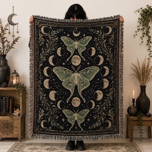 May include: A rectangular woven throw blanket featuring a black background with three large green luna moths centered vertically. The design includes a circular lunar phase cycle surrounding the moths, accented by stars and leafy botanical borders.