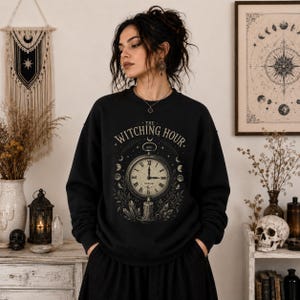 May include: A black crewneck sweatshirt featuring a graphic print of a vintage pocket watch set to 3:00 AM, surrounded by moon phases, crystals, and foliage. The text "THE WITCHING HOUR" is arched above the clock face in a serif font.