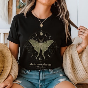 May include: A black short-sleeve t-shirt featuring a central graphic of a pale green moth surrounded by a circular arrangement of moon phases and botanical elements. The text "Metamorphosis by Moonlight" is printed in gold below the moth.