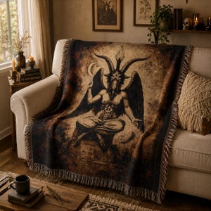 Könnte beinhalten: Eine gewebte Wohndecke mit einer dunklen, sepiafarbenen Illustration einer geflügelten, gehörnten Figur mit einem Pentagramm auf der Stirn, die in meditativer Haltung sitzt. Die Decke hat einen Fransenrand und eine abgenutzte Vintage-Optik.