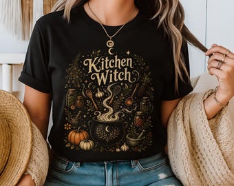 Op de afbeelding: Zwart t-shirt met een gedetailleerde illustratie met een ketel, pompoenen en diverse keukengerei. De tekst "Kitchen Witch" wordt weergegeven in een decoratief lettertype, omringd door kruiden en specerijen. Een halve maan is ook zichtbaar.