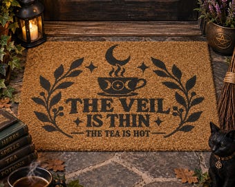 Op de afbeelding: Een rechthoekige deurmat van bruine kokosvezel met een zwarte afbeelding van een maansikkel boven een theekopje, geflankeerd door bladeren. De tekst THE VEIL IS THIN en THE TEA IS HOT staat in een vetgedrukt, donker lettertype op de mat.