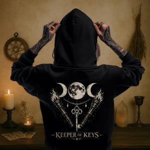 May include: Black hooded sweatshirt with a graphic featuring a full moon, crescent moons, torches, and a key. The text "KEEPER OF KEYS" is printed below the graphic. The sweatshirt is worn by a person with tattooed arms.
