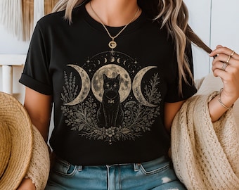 Könnte beinhalten: Ein schwarzes Kurzarm-T-Shirt mit einem Grafikdruck einer schwarzen Katze, die vor einem Vollmond sitzt, flankiert von zwei Mondsicheln und botanischen Elementen. Das Design enthält eine Reihe von Mondphasen, die sich oben wölben.