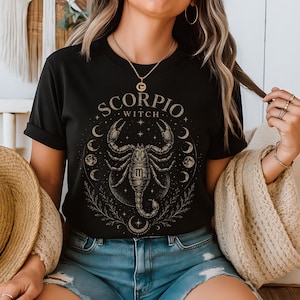 Op de afbeelding: Zwart T-shirt met een gedetailleerde schorpioenafbeelding en de tekst "SCORPIO WITCH". Het ontwerp bevat halve manen en sterren. Het shirt is een T-shirt met korte mouwen en een ronde hals.
