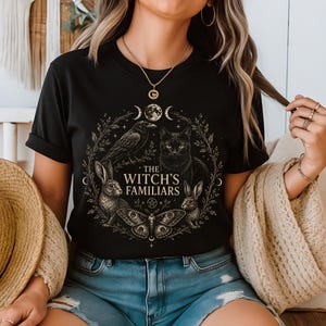 Może przedstawiać: Czarny t-shirt z krótkim rękawem z okrągłym nadrukiem graficznym w odcieniach złota i bieli. Wzór przedstawia kruka, kota, dwa króliki i ćmę w otoczeniu liści, księżyca oraz napisu THE WITCH'S FAMILIARS umieszczonego na środku grafiki.