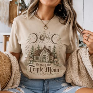 Könnte beinhalten: Ein beigefarbenes T-Shirt mit einem Grafikdruck eines Häuschens, umgeben von Tannen und Pilzen, gekrönt von drei Mondphasen. Der Text Beneath the Triple Moon ist unter dem Haus in einer dekorativen Schriftart mit floralen Akzenten gedruckt.