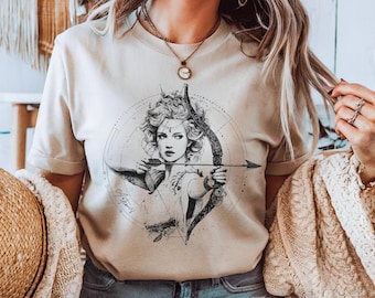 Könnte beinhalten: Beiges T-Shirt mit einer detaillierten Schwarz-Weiß-Illustration einer Frau mit Pfeil und Bogen. Das Design beinhaltet ein kreisförmiges Element mit gepunkteten Linien und dem Wort "Artemis". Das Shirt hat kurze Ärmel.