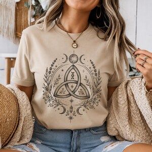 Può includere: Una t-shirt beige a maniche corte con una stampa grafica nera sul petto. Il design mostra un nodo triquetra centrale circondato da elementi botanici, fasi lunari, stelle e un cristallo, creando un dettagliato motivo circolare.