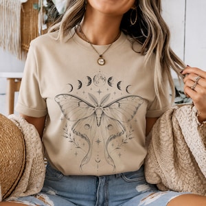 Op de afbeelding: Een beige t-shirt met korte mouwen en een zwarte inktillustratie van een grote nachtvlinder op de borst. Boven de vlinder staat een horizontale rij maanfasen en het ontwerp is versierd met kleine sterretjes en verfijnde botanische takjes.