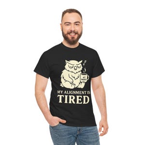 Op de afbeelding: Zwart T-shirt met een crèmekleurige uilafbeelding die een koffiemok vasthoudt met "+1 Energy" erop. De tekst "MY ALIGNMENT IS: TIRED" staat onder de uil. Het T-shirt wordt door een persoon gedragen.
