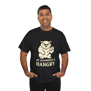 Può includere: T-shirt nero con una grafica di gufo color crema che tiene un piccolo oggetto. Il testo "MY ALIGNMENT IS: HANGRY" è stampato sotto. La maglietta è un capo casual.
