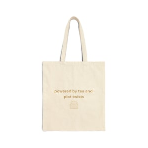 May include: A cream-coloured tote bag with long handles. The bag has the text "powered by tea and plot twists" in gold, and a small illustration of an open book. The bag is made of a sturdy fabric, ideal for carrying books and other items.